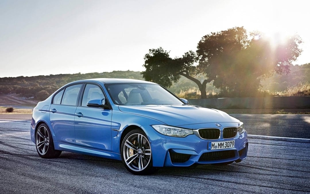 Yas Marina Blue BMW M3 sedan on a racetrack with a bright sun flare and green hills in the background.