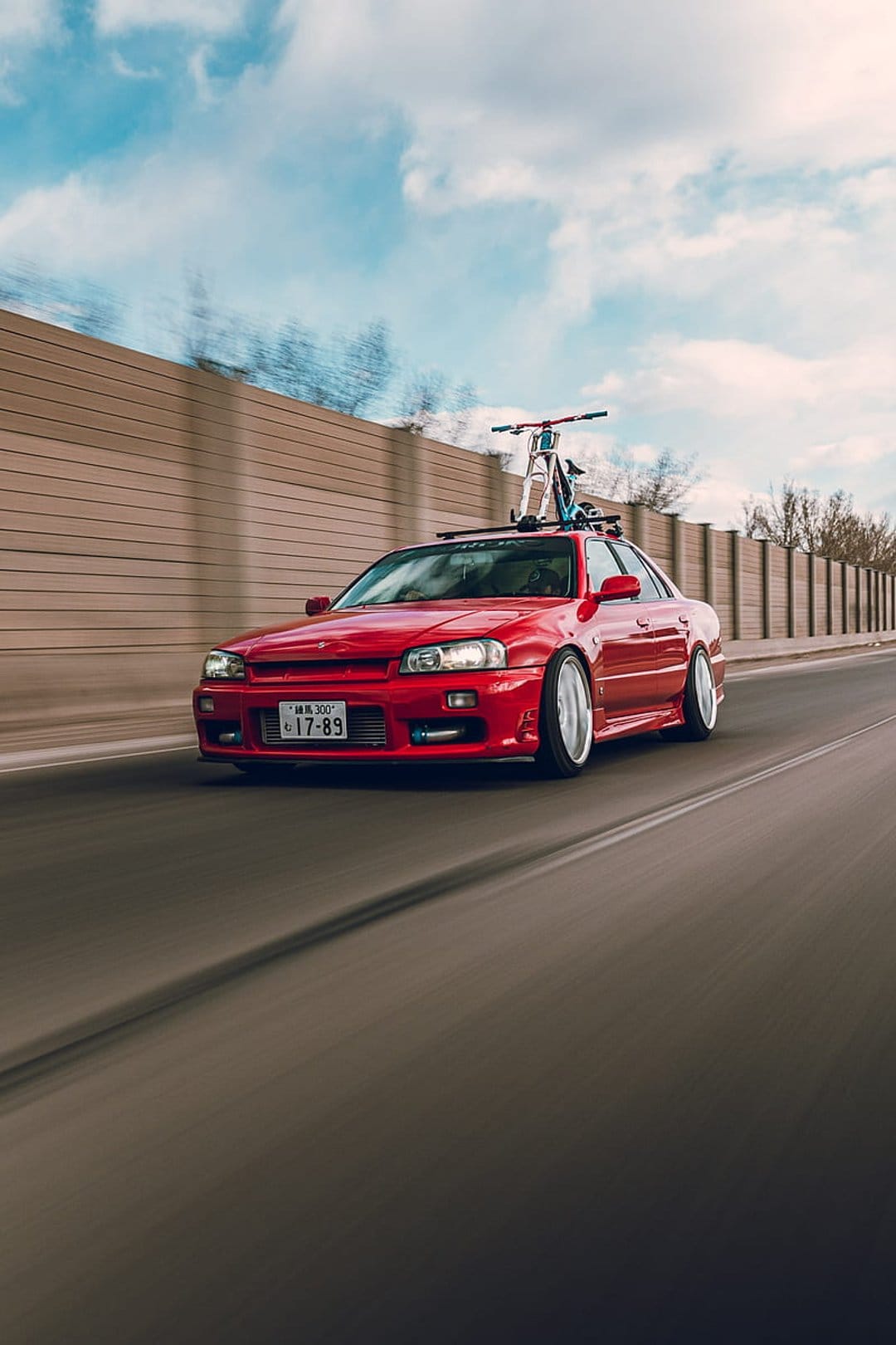 Red Nissan Skyline R34 sedan with a bicycle on its roof rack driving past a blurred highway sound barrier.