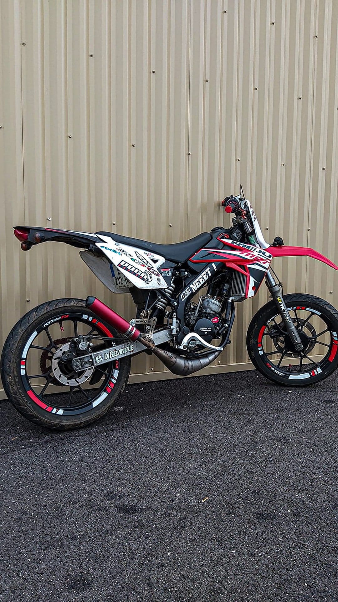 Red and white supermoto motorcycle with custom decals and red rims parked against a corrugated beige metal wall.