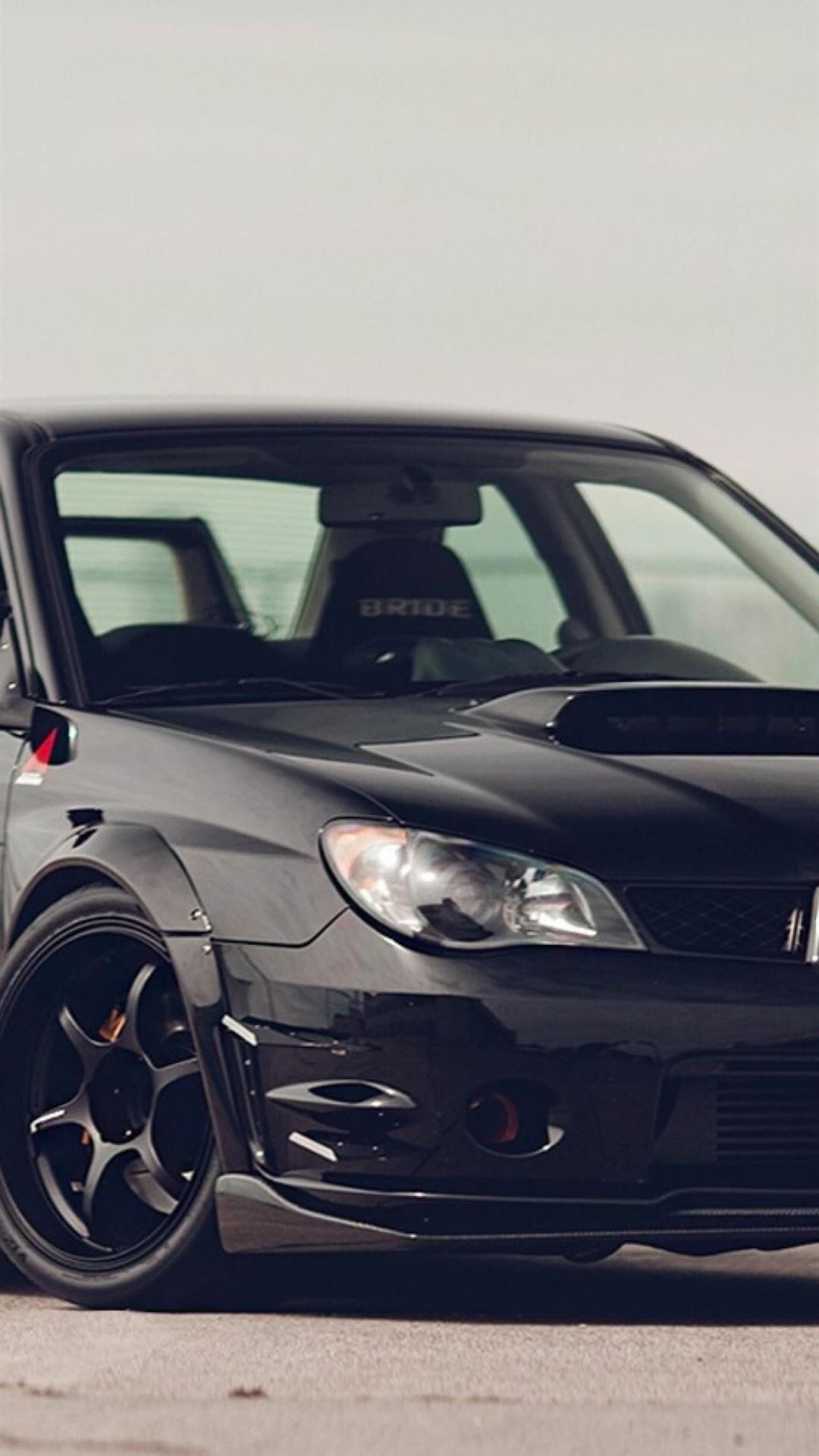 Close-up of a black Subaru WRX STI with widebody fenders, black wheels, and a hood scoop in soft daylight.