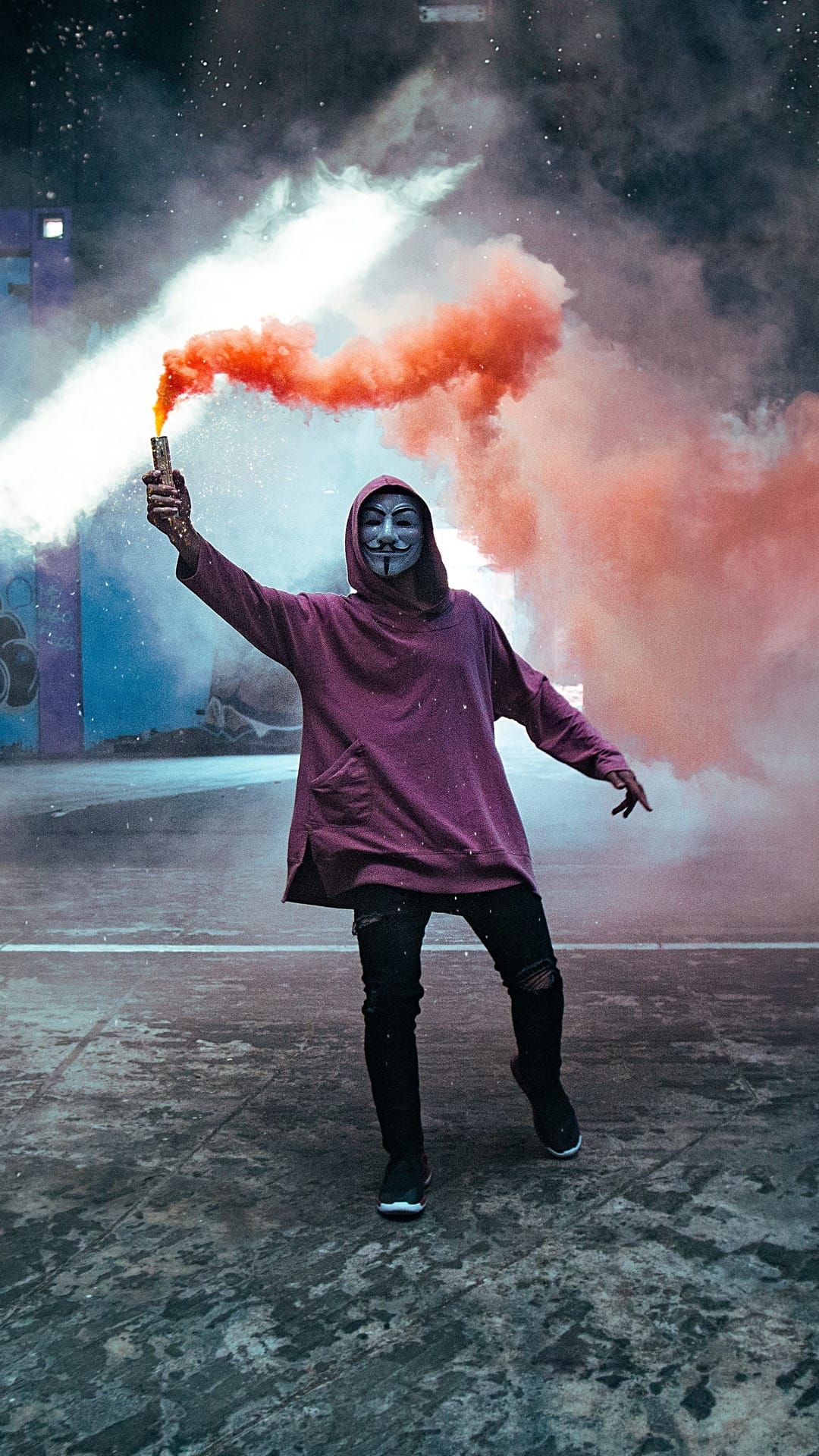 Masked person in a maroon hoodie holds a canister emitting thick orange smoke against a city building background.