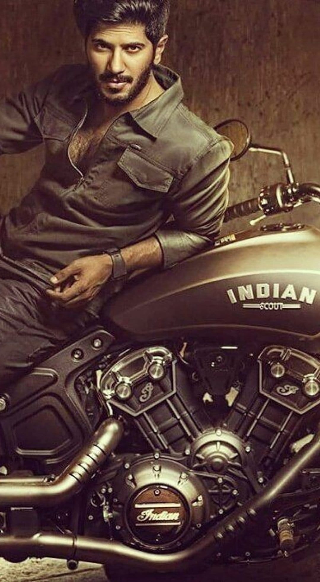 Bearded man in a green jacket sits on a brown Indian Scout motorcycle against a textured dark wall background.