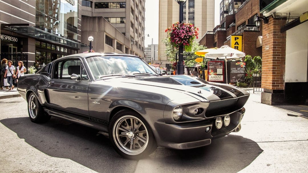 Gray Shelby GT500 Eleanor Mustang with black stripes parked on a sunlit city street with urban buildings.