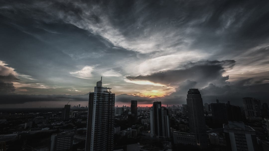 City skyscrapers stand beneath swirling dark clouds and golden sunset light during a dramatic storm.
