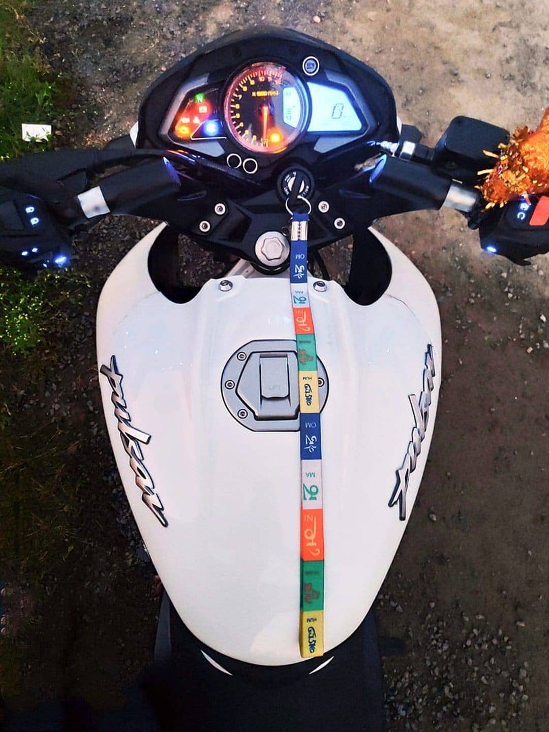 White Bajaj Pulsar motorcycle dashboard and fuel tank with a colorful lanyard viewed from a top-down angle.