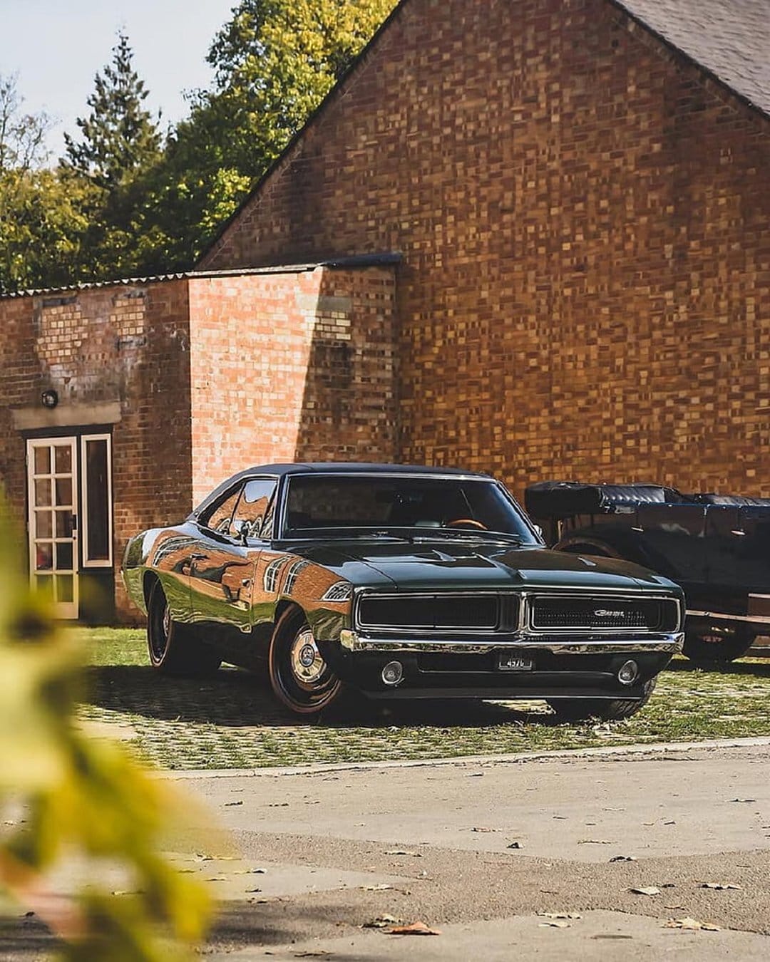 1969 Dodge Charger R/T in dark green parked on pavement near a red brick building with sunlight reflections.