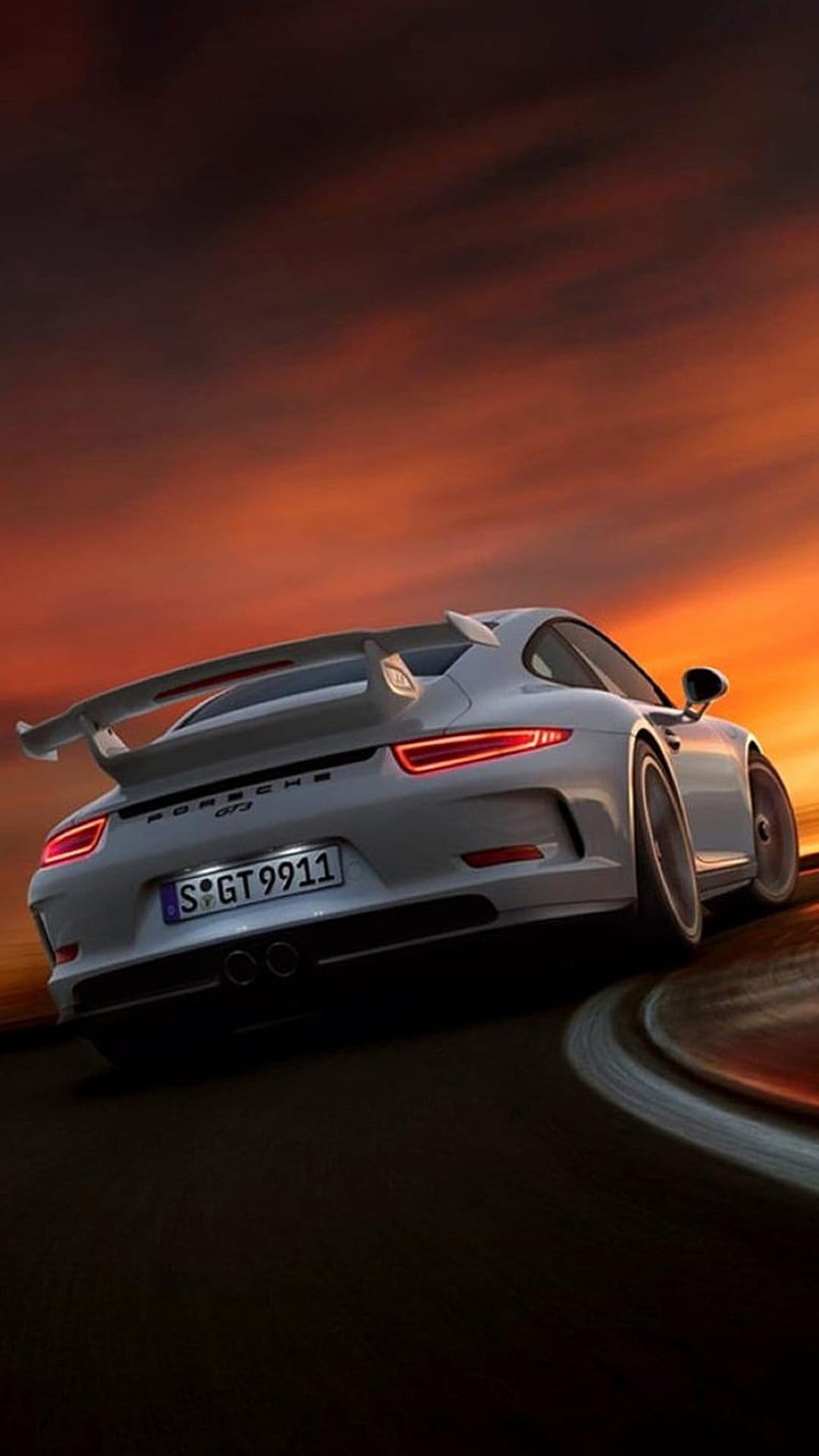 White Porsche 911 GT3 with glowing red taillights corners on a race track beneath a vibrant orange sunset sky.
