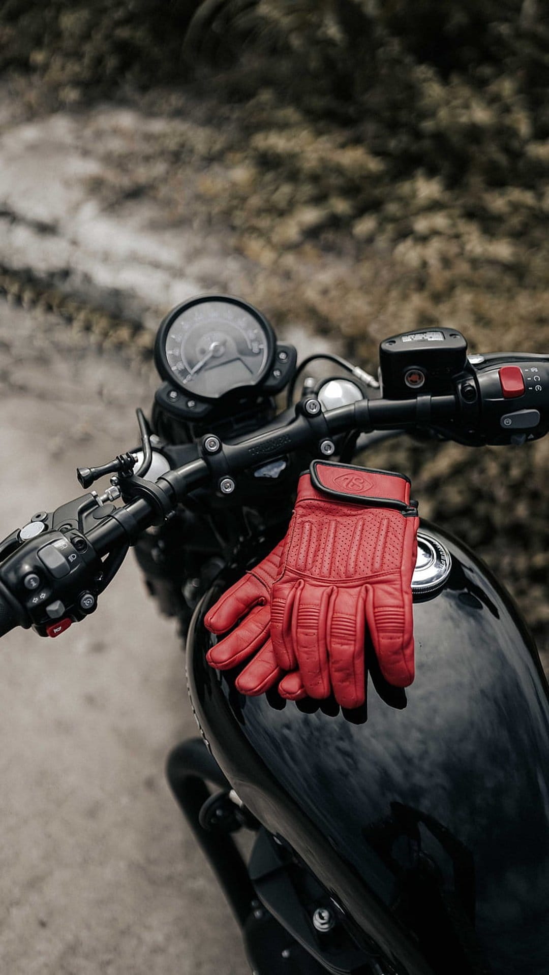 Red leather motorcycle gloves lie on a glossy black tank with a vintage speedometer in the soft-focus background.