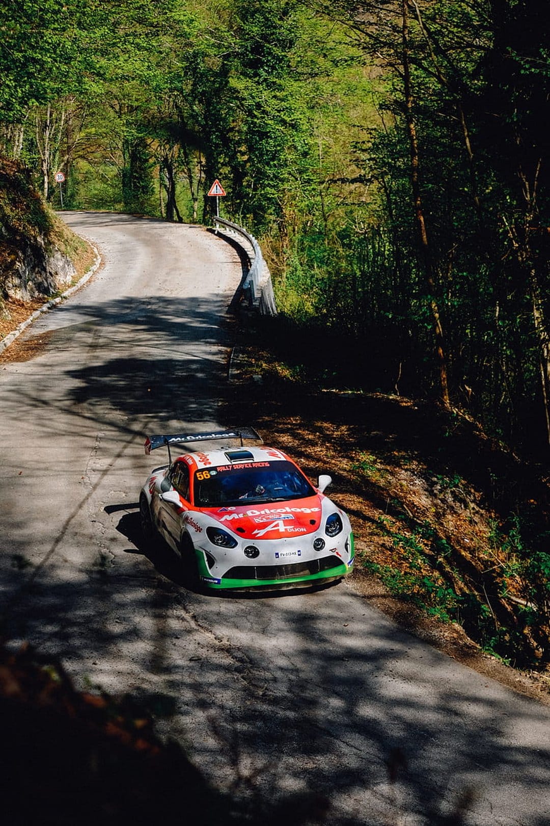 Alpine A110 rally car with red and green livery drifting on a winding asphalt road through a sunlit dense forest.