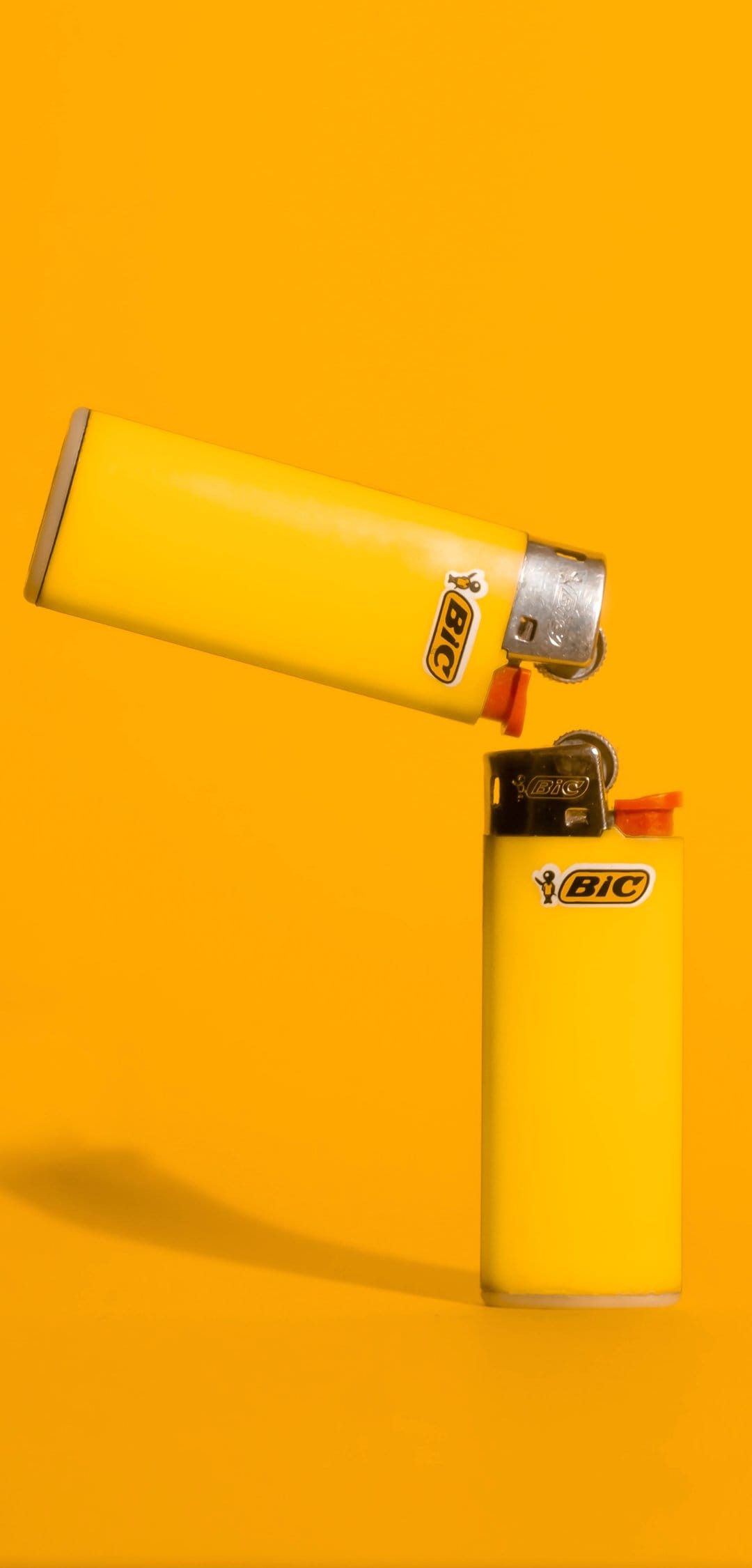 Two yellow butane lighters with chrome caps positioned on a vibrant orange background with soft shadows.