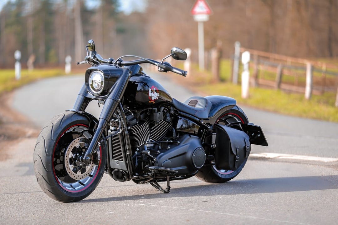 Custom black cruiser motorcycle with fat tires and a leather saddlebag on an asphalt road during daylight.