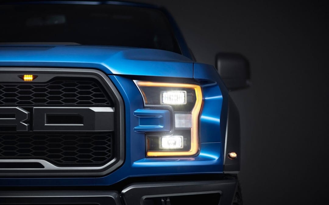 Close-up of a blue Ford Raptor truck's LED headlight and black honeycomb grille under dramatic studio lighting.