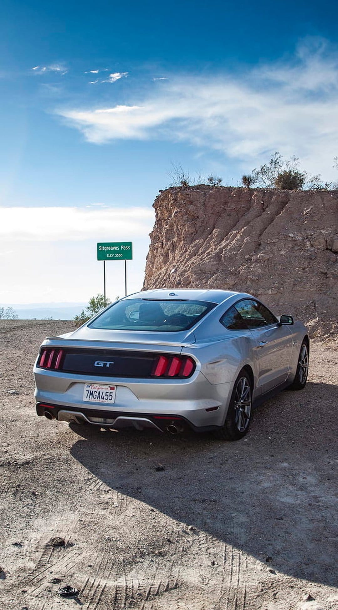 Silver Ford Mustang GT parked on a dusty road beside a rocky cliff and Sitgreaves Pass sign under a blue sky.