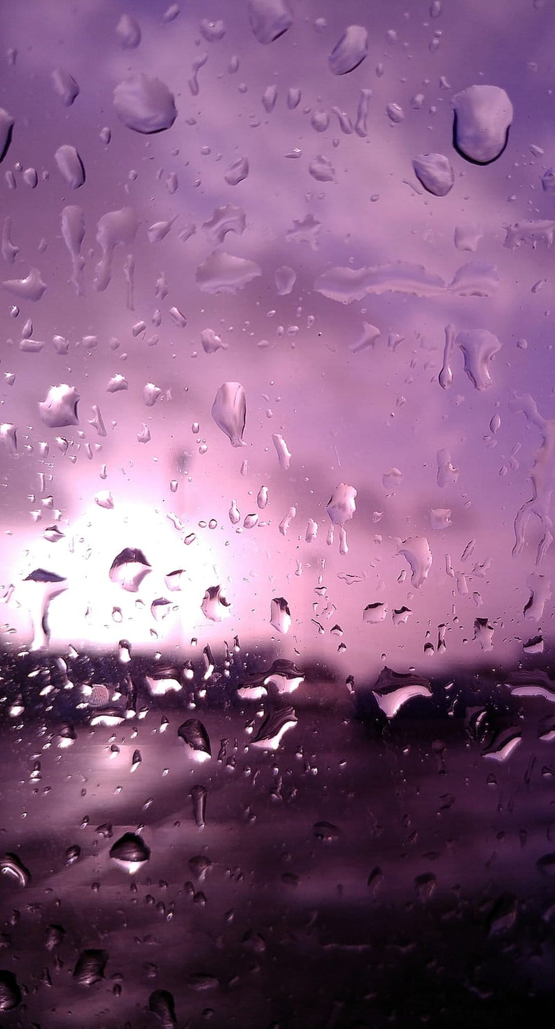 Purple water droplets on a windowpane refract the glowing violet and pink hues of a twilight sky at dusk.