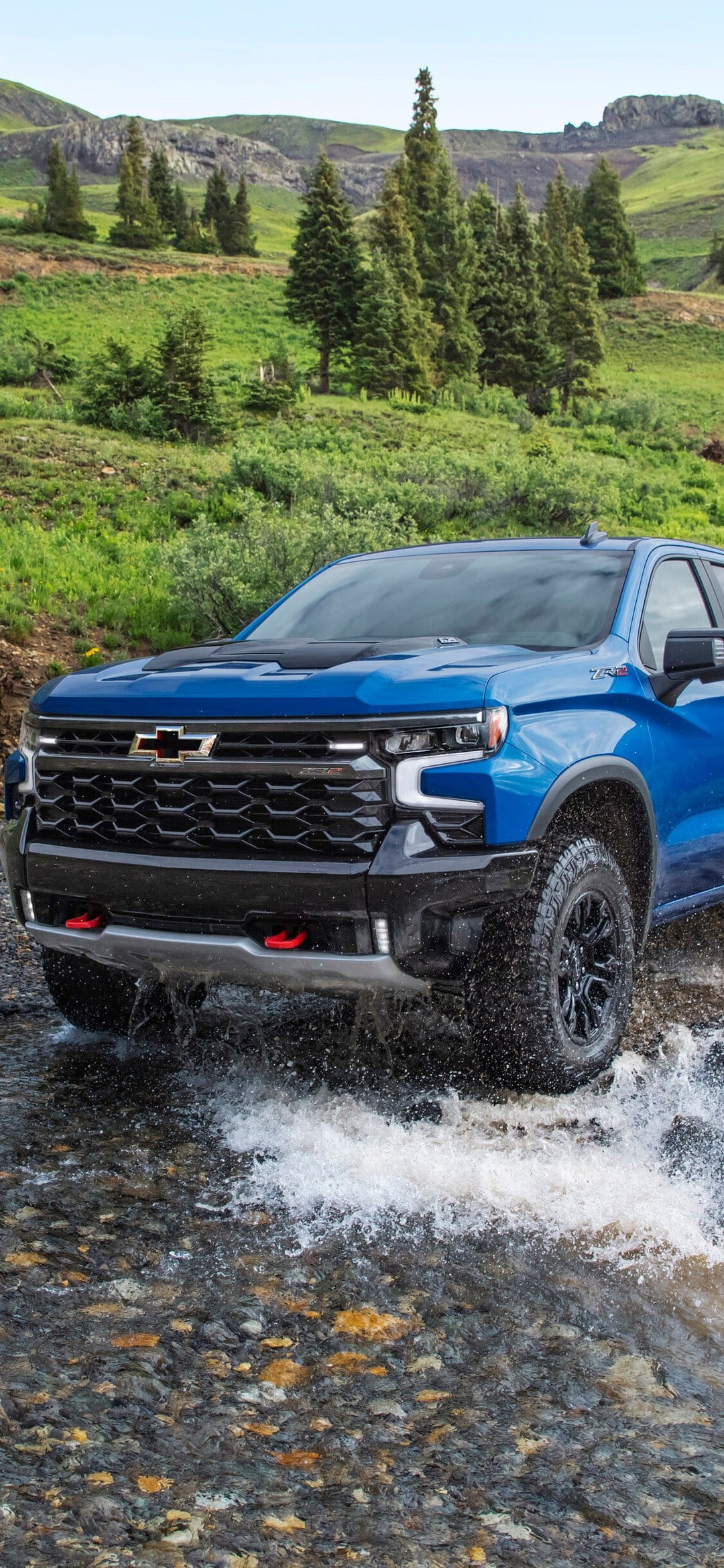 Blue Chevrolet truck splashing through a rocky mountain river with green pine trees in the background.