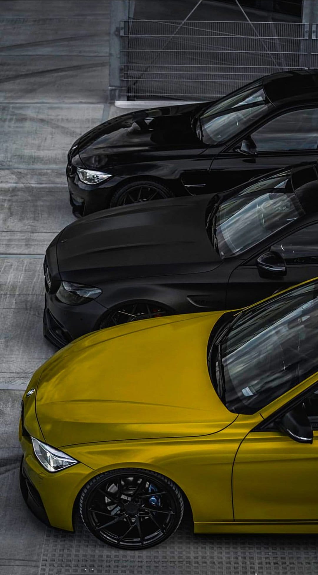 Yellow BMW followed by two matte black cars parked in an industrial garage with concrete floors and metal grates.