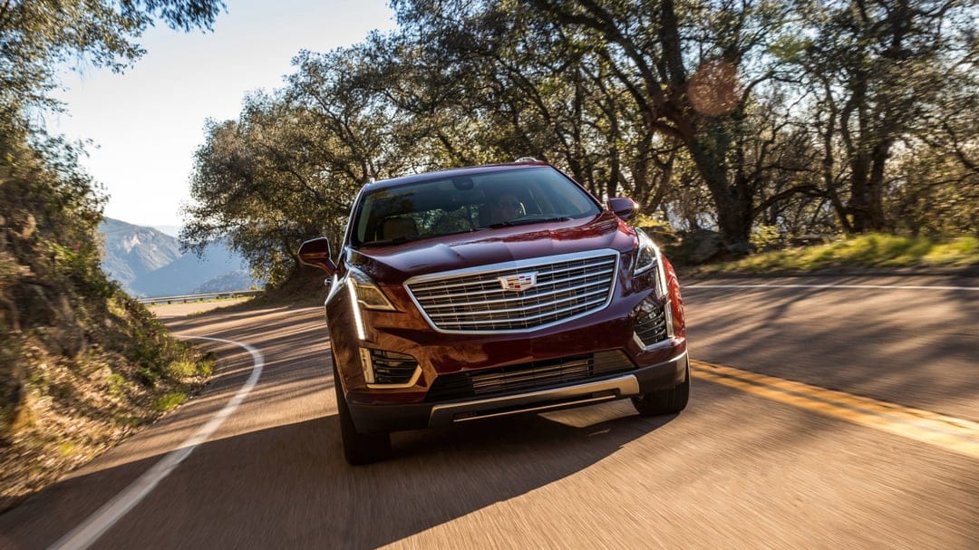 Maroon Cadillac SUV driving along a sunlit winding road through a forest with distant blue mountains.