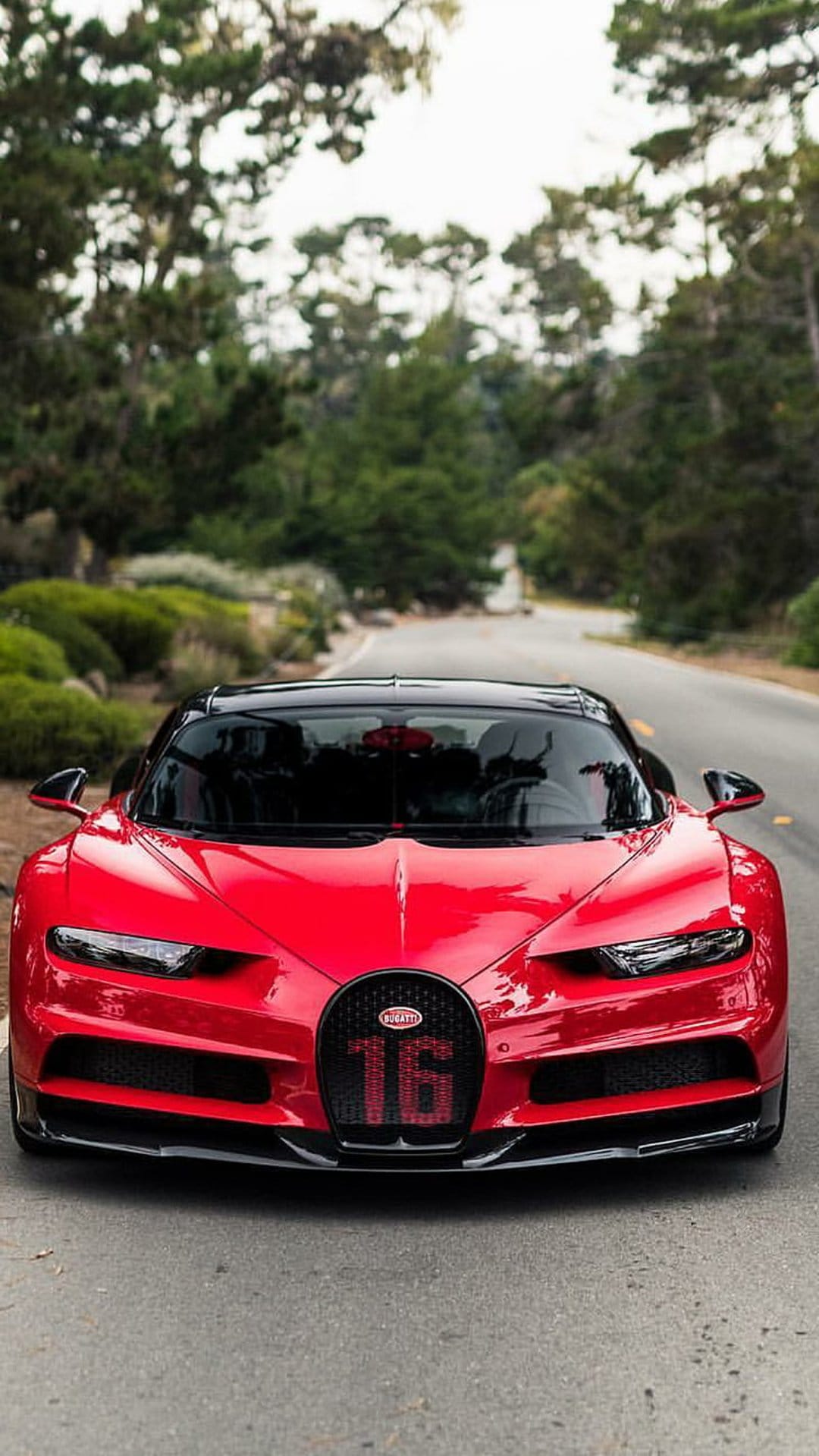Red Bugatti Chiron 16 hypercar with a glowing 16 grille parked on a scenic road among lush green trees.