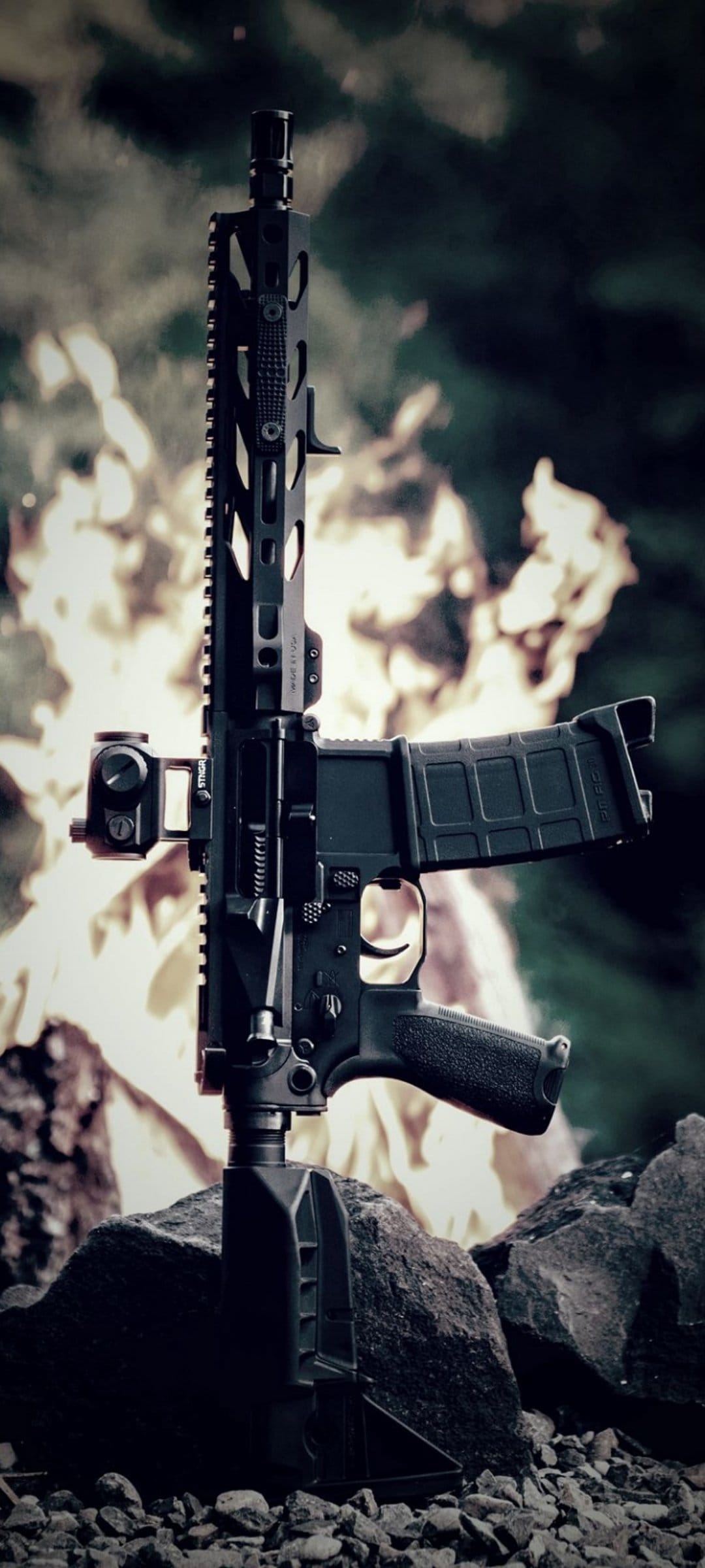 AR-15 rifle with a red dot sight sits on grey rocks with orange flames and green leaves in the background.