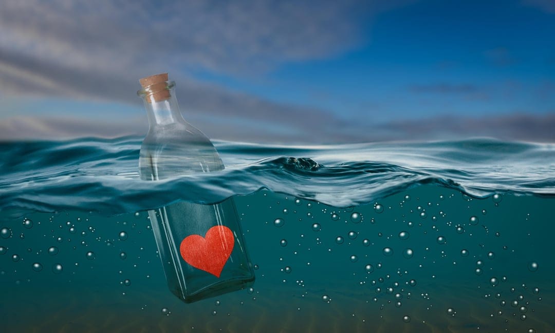 Red heart inside a corked glass bottle floating in blue ocean water with sunlight and surface bubbles.