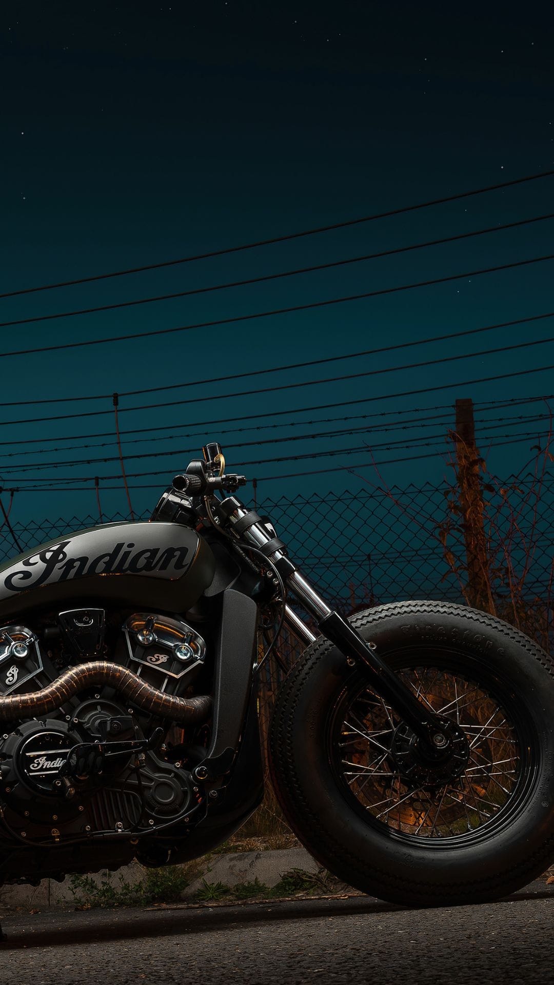 Custom Indian motorcycle with a matte tank and thick tires parked against a teal night sky and wire fence.