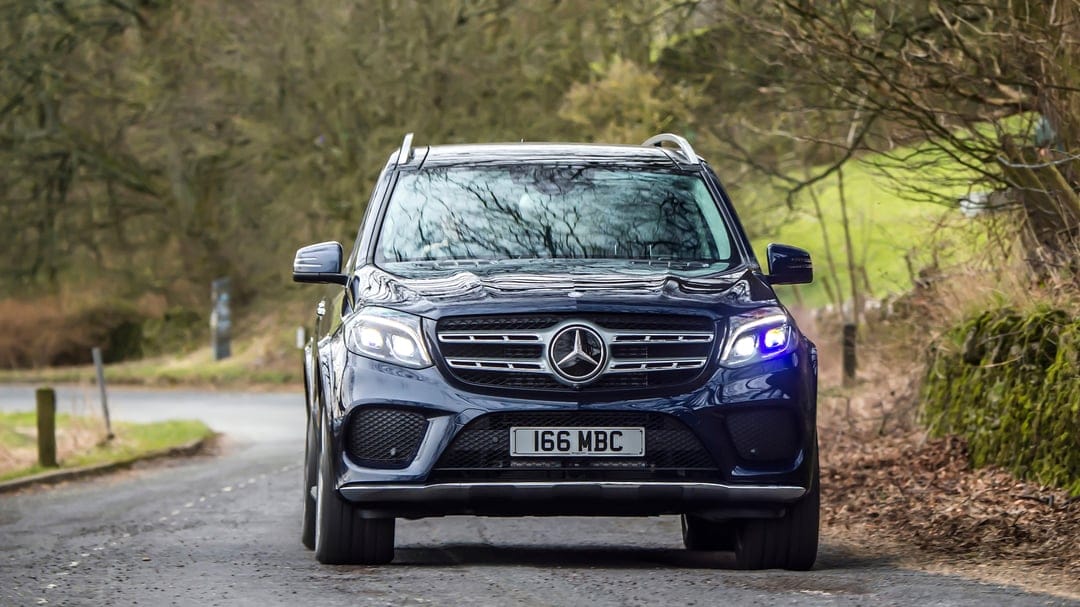Navy blue Mercedes-Benz GLS SUV driving forward on a rural asphalt road between leafless trees and green fields.