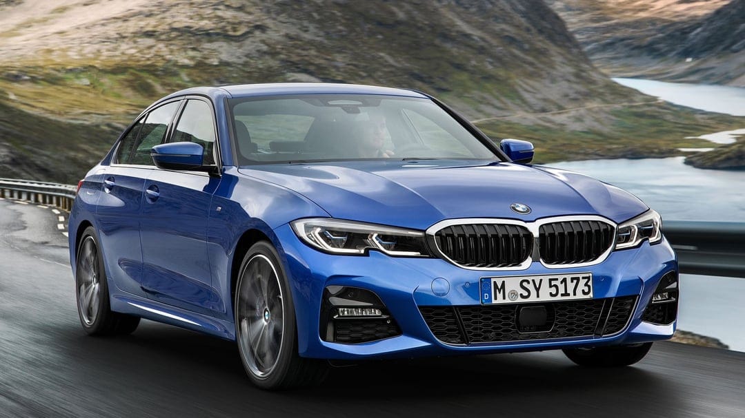 Blue BMW 3 Series sedan driving on a winding mountain road with a scenic lake and rocky hills in the background.