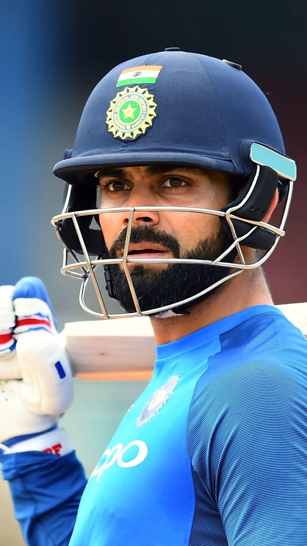 Virat Kohli wearing a blue Indian cricket jersey and protective helmet with a focused expression during a game.