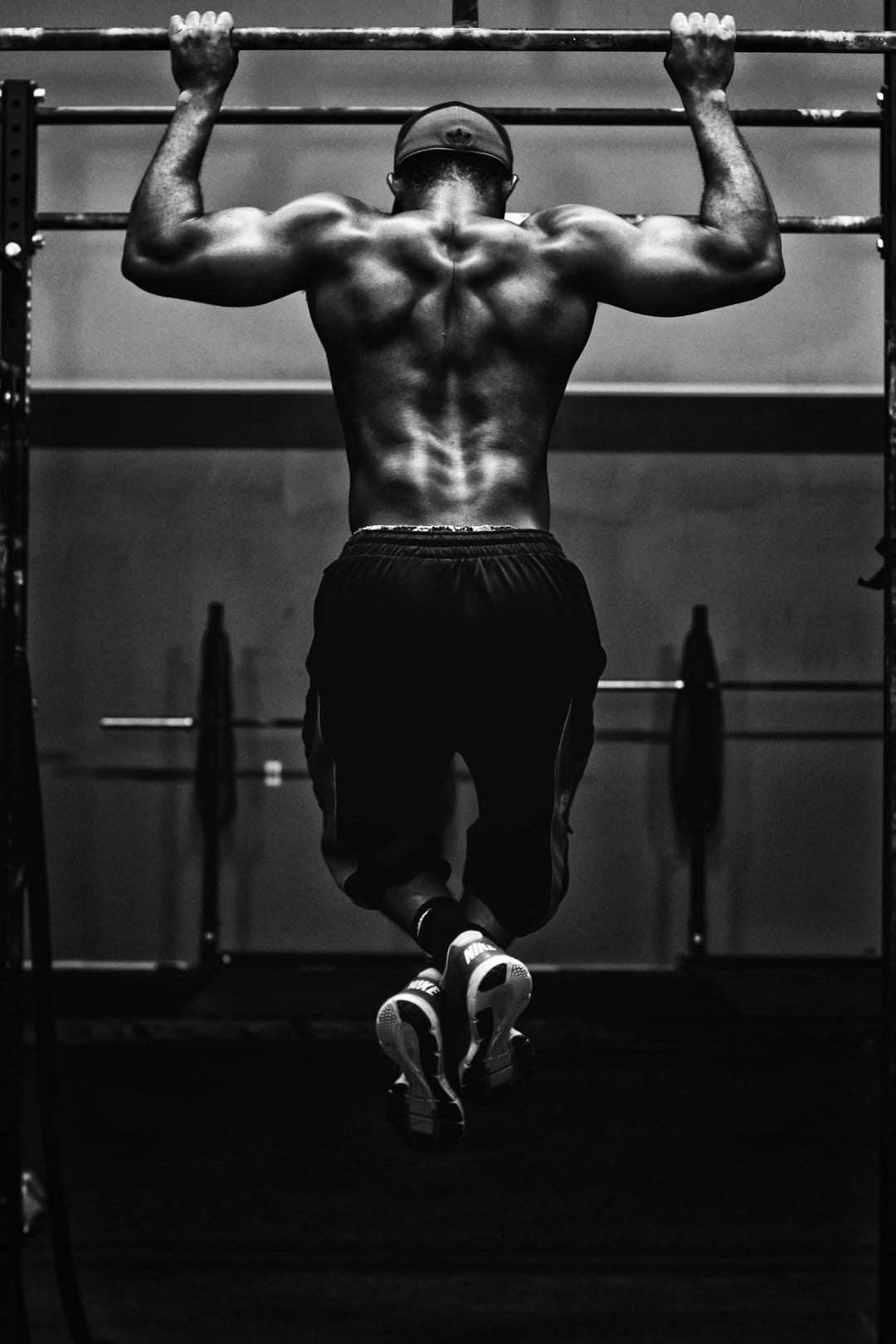 Muscular man performing pull-ups with defined back muscles in a high-contrast black and white gym setting.