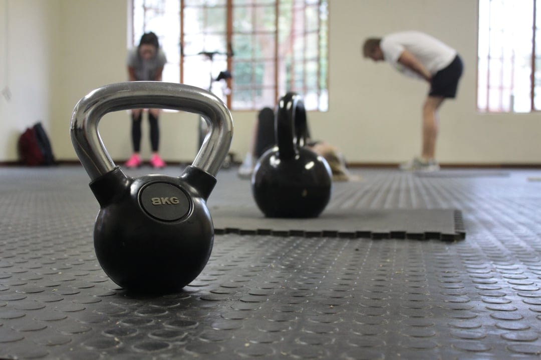 Polished metal kettlebell sits in the foreground of a bright gym with blurred figures exercising by large windows.