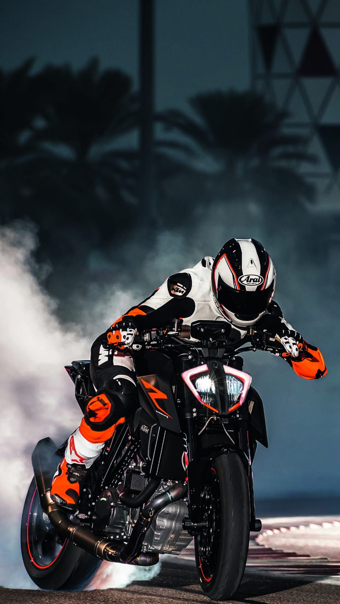 Motorcyclist in an orange suit leans on a black bike while thick white smoke billows from a spinning rear tire.