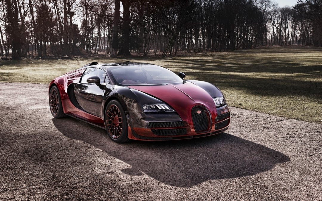 Red and black carbon fiber Bugatti Veyron parked on a gravel path in a wooded park area under soft light.