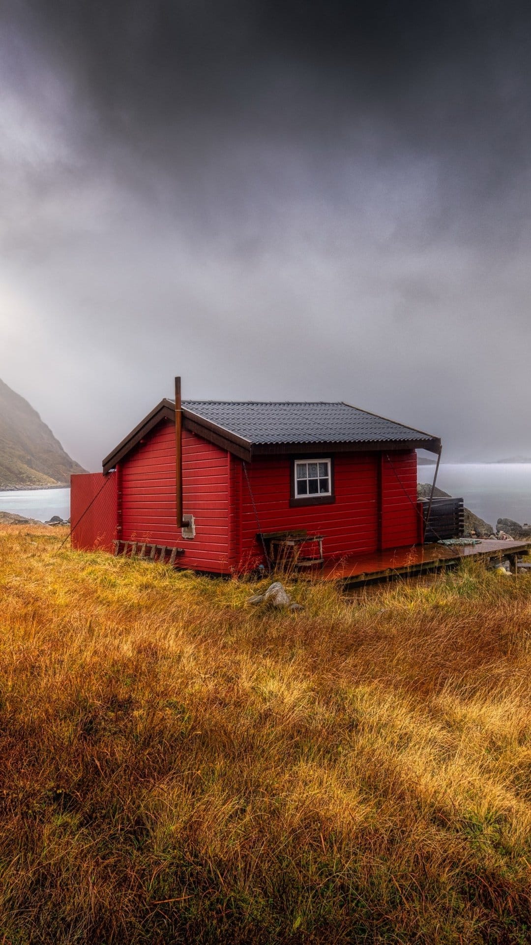 Red wooden cabin stands on golden coastal grass under a heavy overcast sky near the grey northern ocean waves.