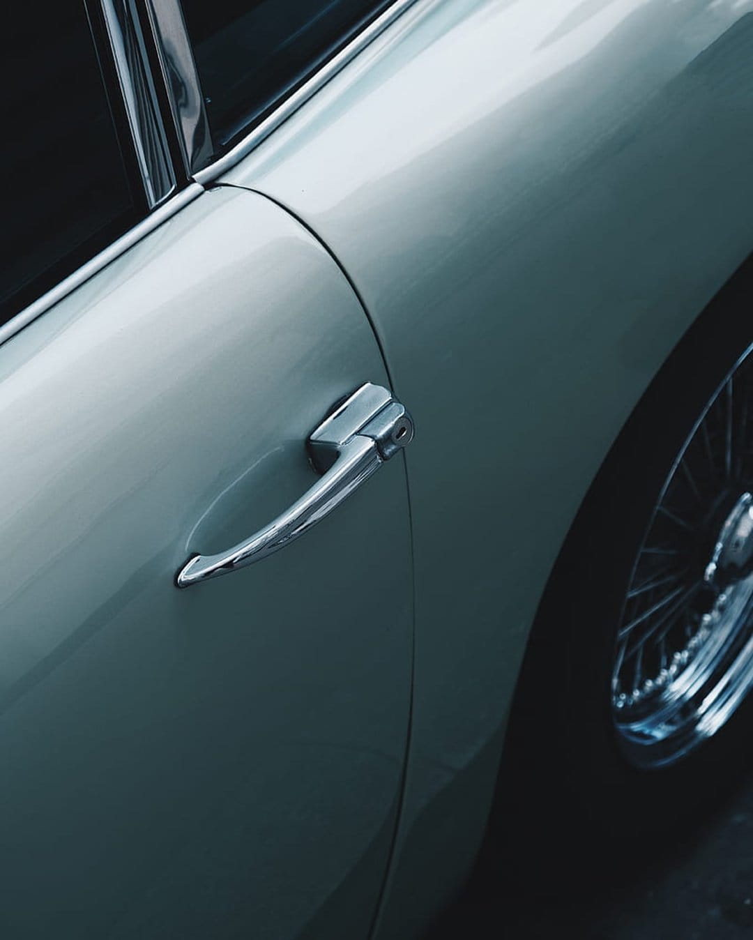 Chrome door handle and a wire-spoke wheel on a vintage car with muted bodywork and reflective metallic finishes.