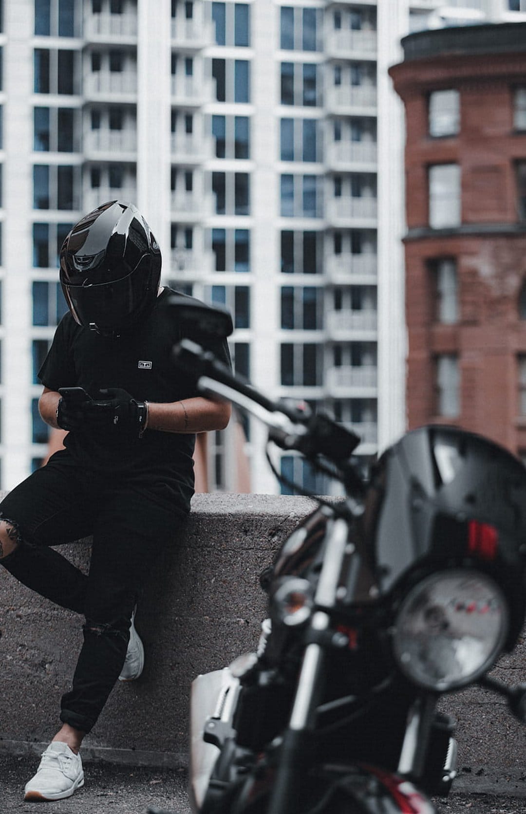 Motorcyclist in a black helmet and gloves looks at a smartphone while sitting near a blurred motorcycle in a city.