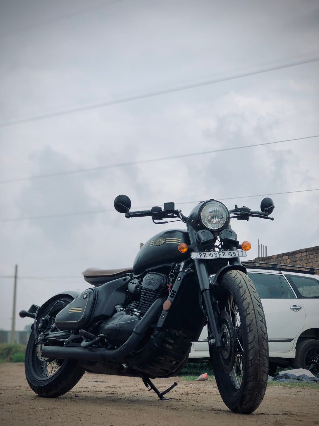 Black Jawa motorcycle with a brown seat parked on a dusty street under an overcast sky with power lines above.