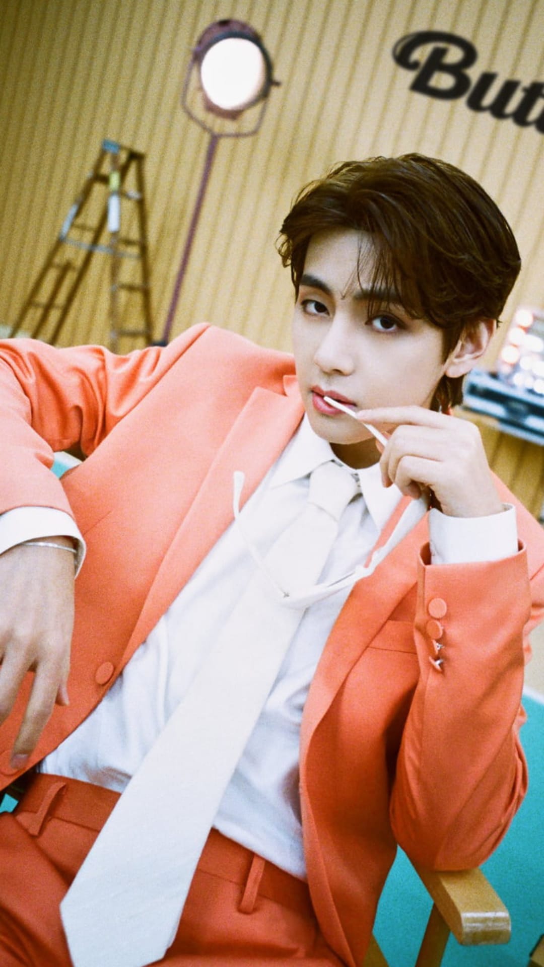 Kim Taehyung in a peach suit holds a lollipop to his lips against a studio backdrop with dramatic lighting.