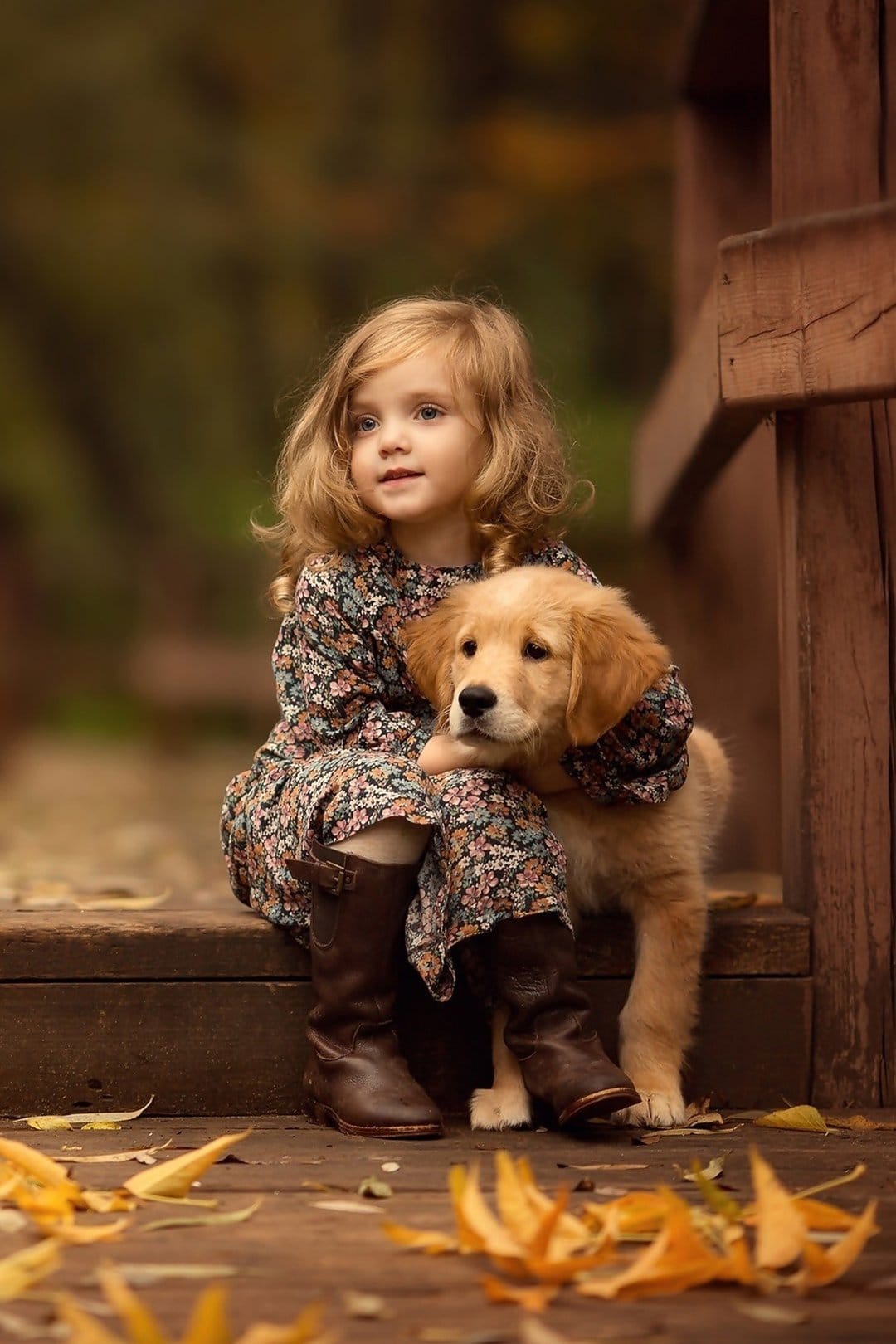 Blonde girl in a floral dress hugs a golden retriever puppy while sitting on a leaf-covered wooden bridge.