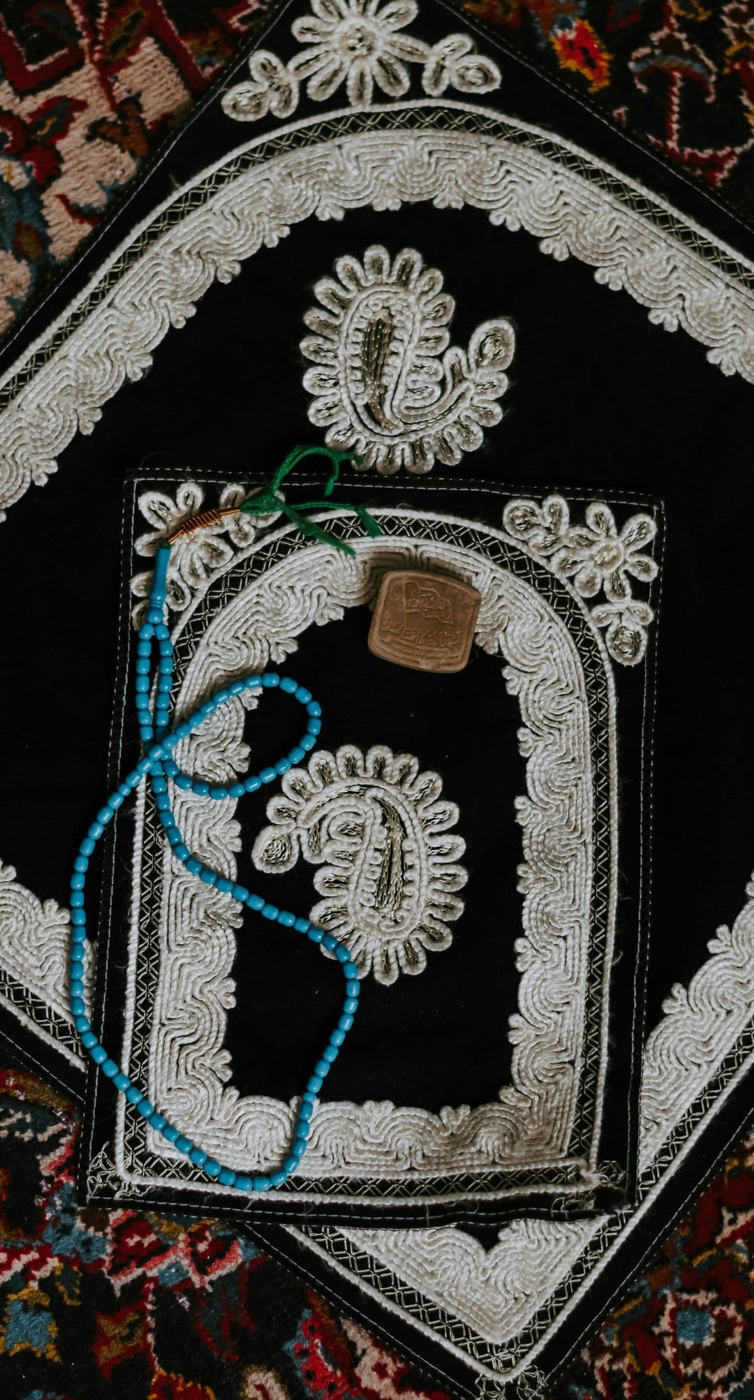 Black prayer rug with white embroidery and blue tasbih beads resting on a patterned carpet for phone display.