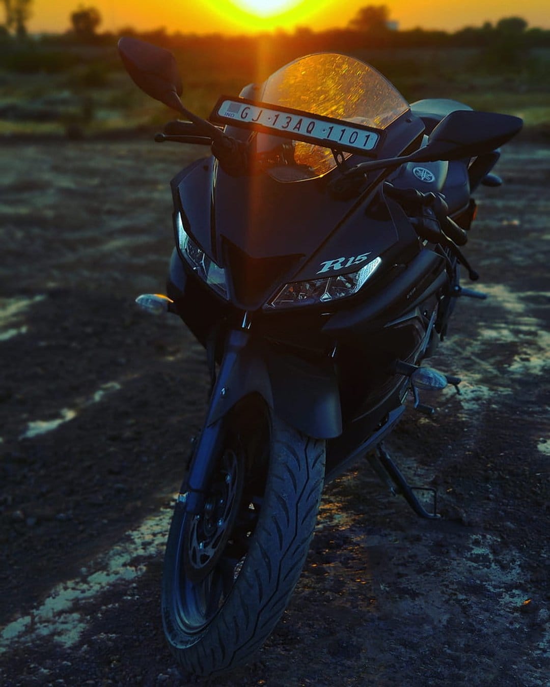 Black Yamaha R15 motorcycle parked on a muddy rural path under a vibrant orange and golden sunset sky.