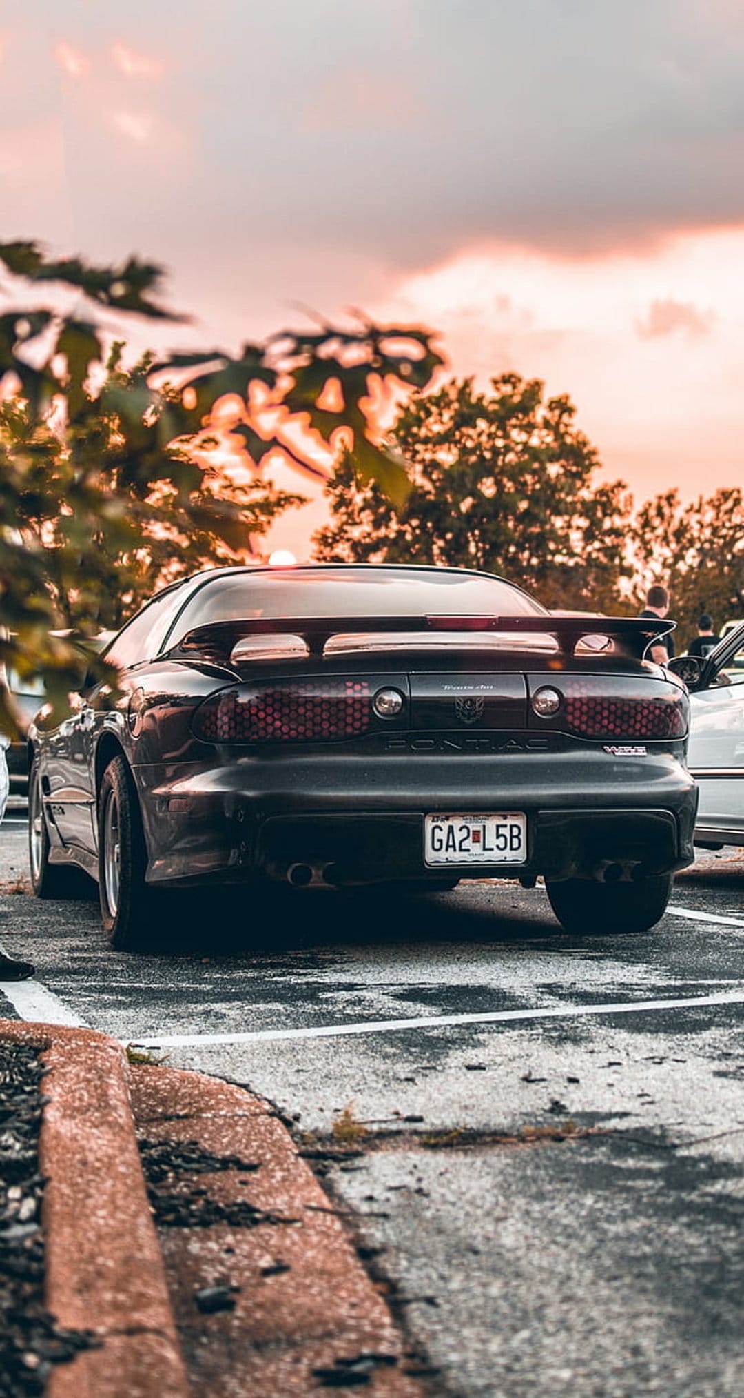 Black Pontiac Firebird rear view with glowing taillights parked under a vibrant orange and pink sunset sky.