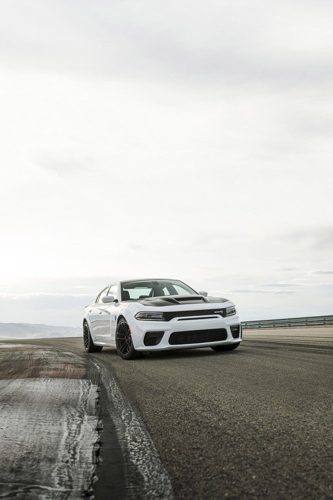 White Dodge Charger SRT Hellcat Widebody with a black hood parked on a textured asphalt racetrack under overcast skies.