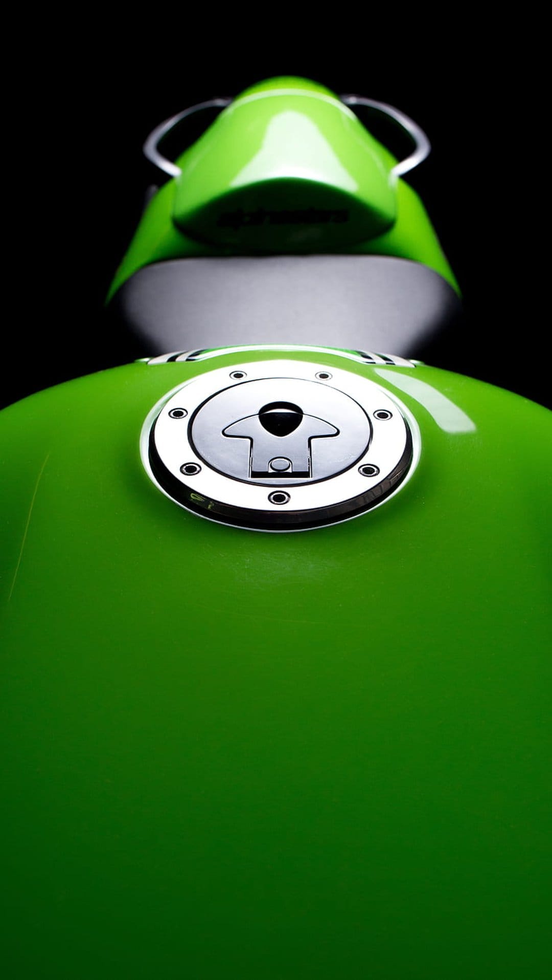 Green motorcycle fuel tank with a polished chrome cap and hex bolts isolated against a dark, shadowy background.