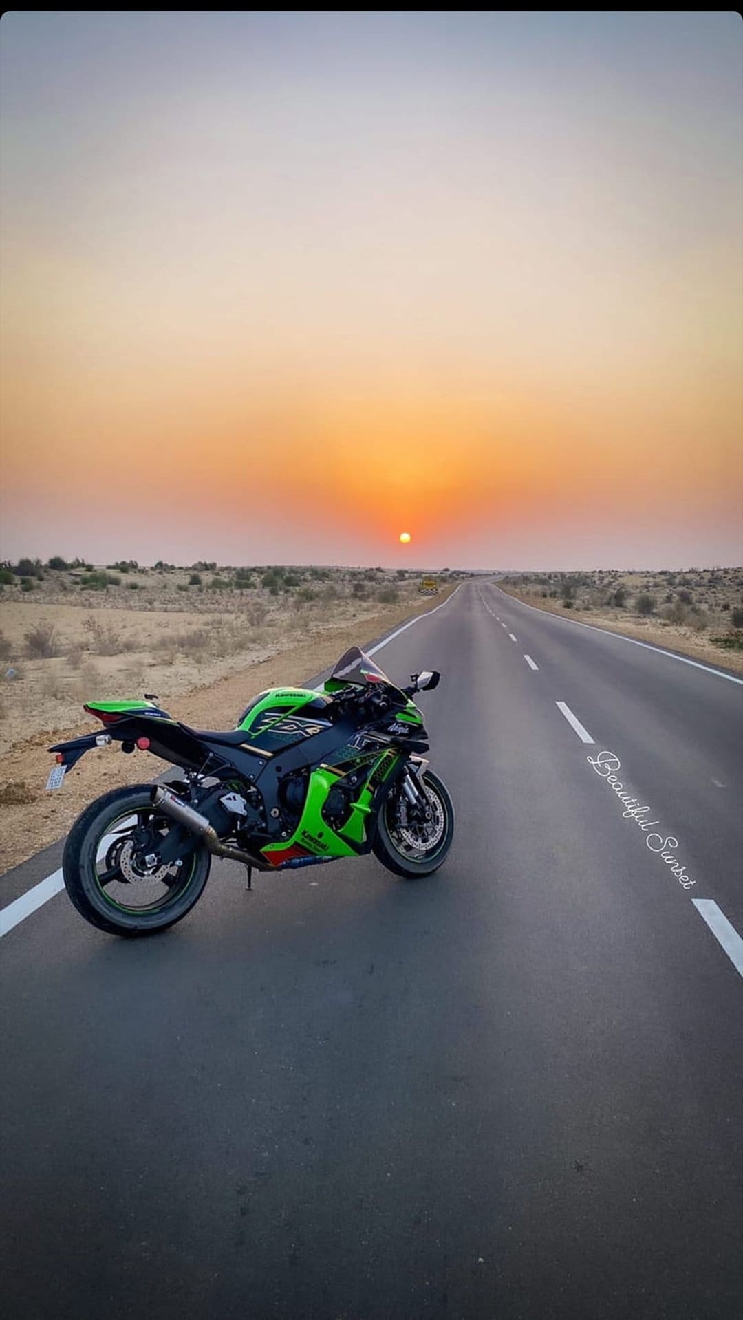 Green sportbike parked on a desert highway shoulder under a vibrant orange sunset with a distant horizon line.