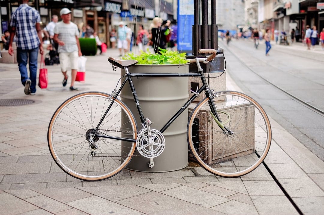 Black vintage bicycle parked by a stone planter with green foliage on a cobblestone street with distant people.