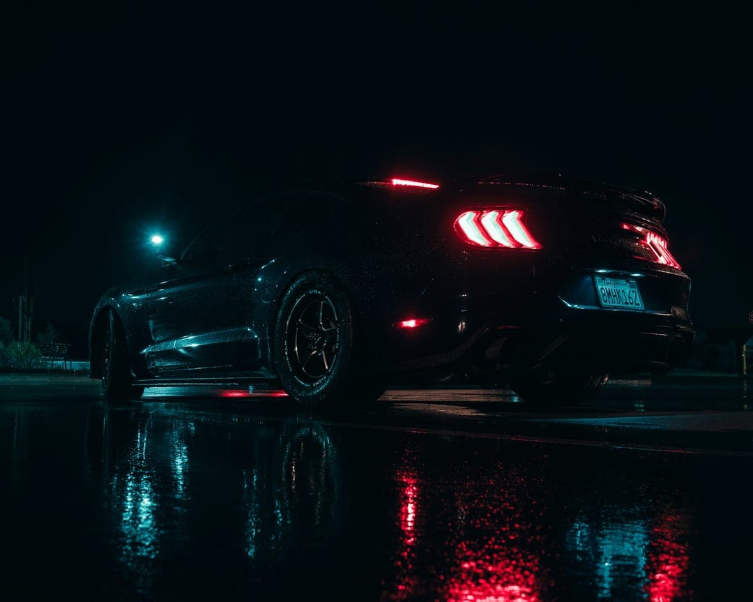Ford Mustang GT rear view on a rain-slicked road at night with glowing red taillights and blue neon reflections.