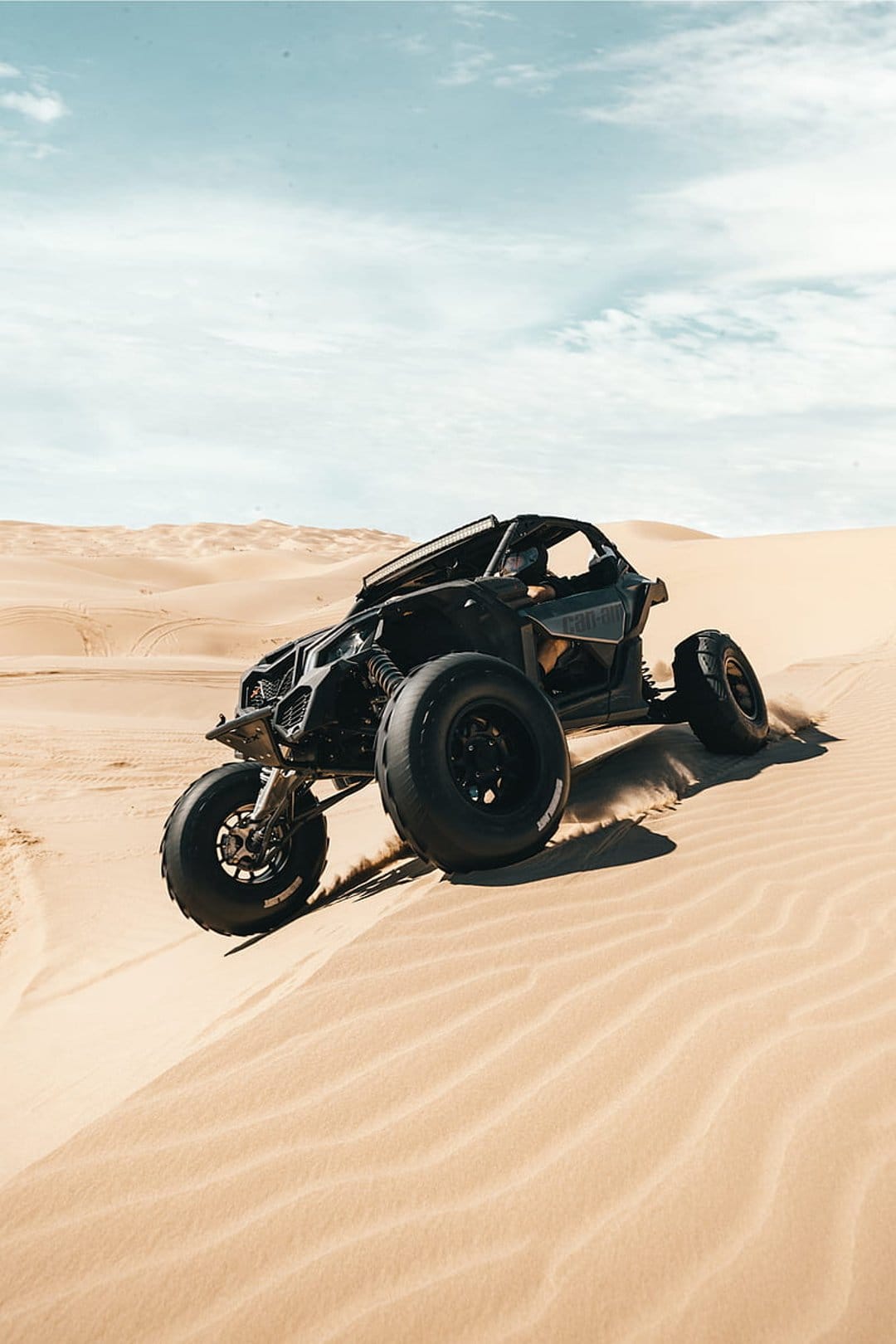 Black dune buggy kicking up sand while driving across desert hills under a blue sky with scattered clouds.