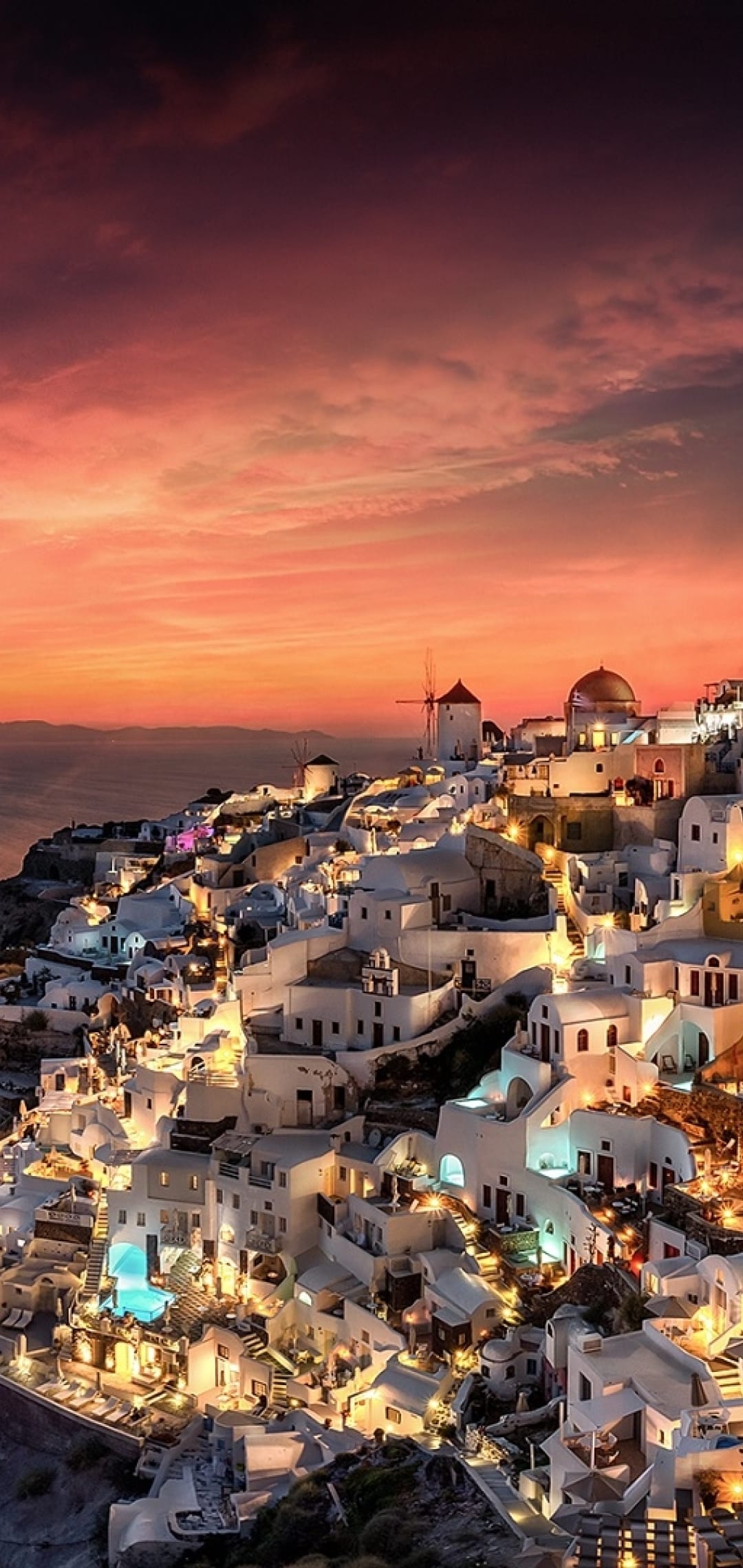 Whitewashed houses of Oia glow with warm lights against a fiery sunset sky above the Aegean Sea at twilight.