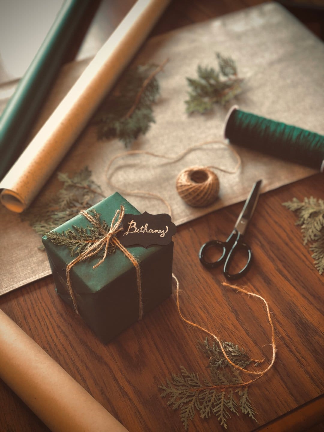Dark green gift box with pine sprigs and Bethany tag on a wooden table surrounded by twine and scissors.