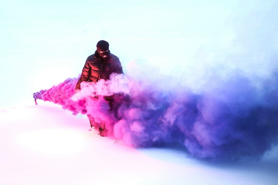 Masked person in a black puffer jacket releases vibrant pink and purple smoke clouds across a snowy white field.